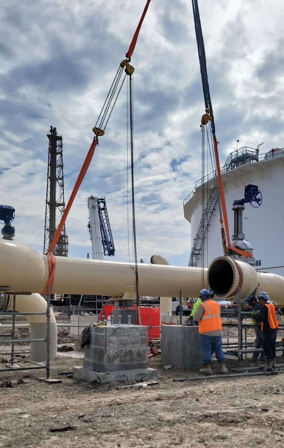 Riggers on a refinery site guiding a large pipe section into place, lifted on slings and chain hoists beneath a crane's hook