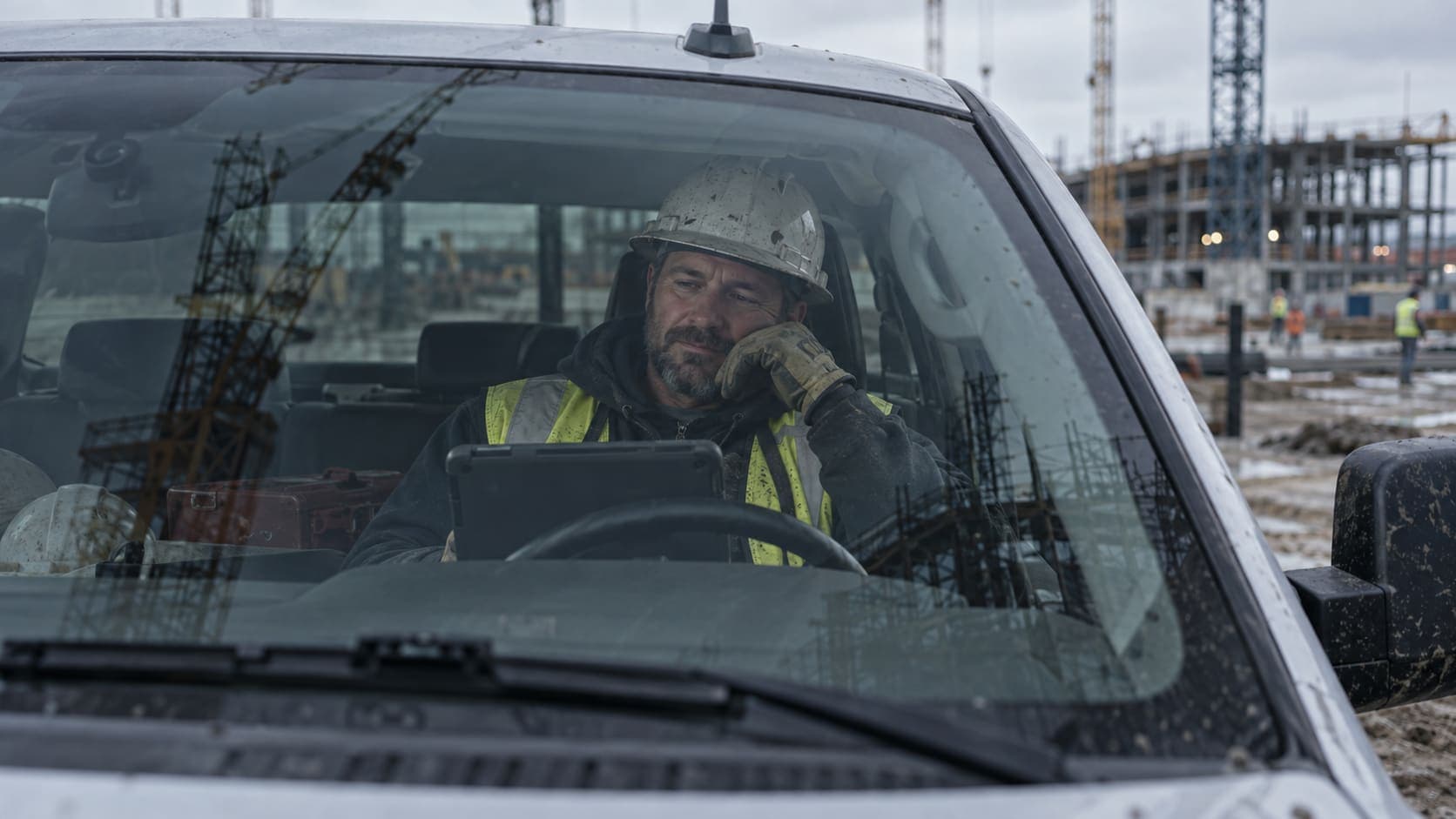 A crane operator sitting in a pickup truck at a job site, reading a tablet during a break